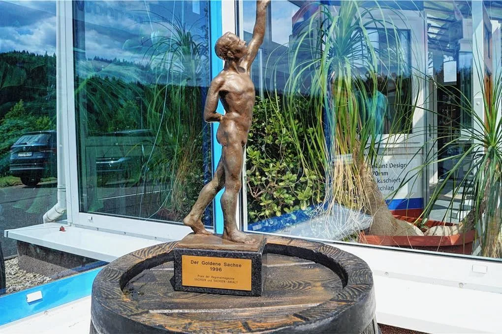 Die Auszeichnung "Der Goldene Sachse 1996", präsentiert auf einem dunklen Holzfass im Schaufenster eines Büros mit Blick auf ein Auto und Pflanzen.
