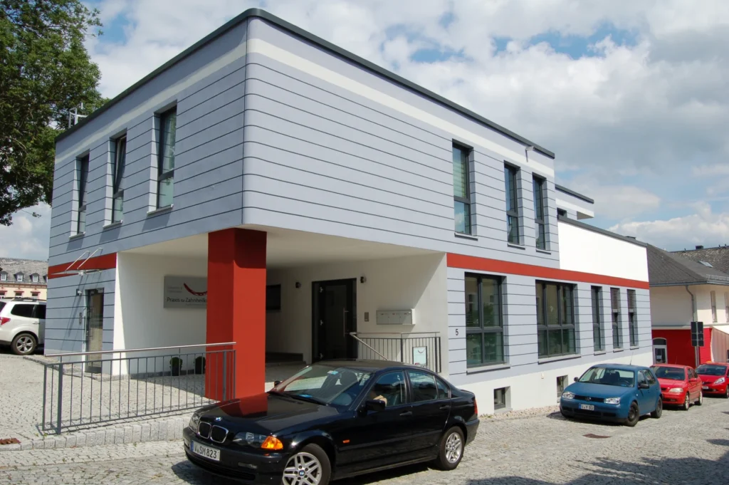 Moderne Zahnarztpraxis in Treuen mit grauer Fassade, roten Akzenten, Aluminiumfenstern und Eingangstür